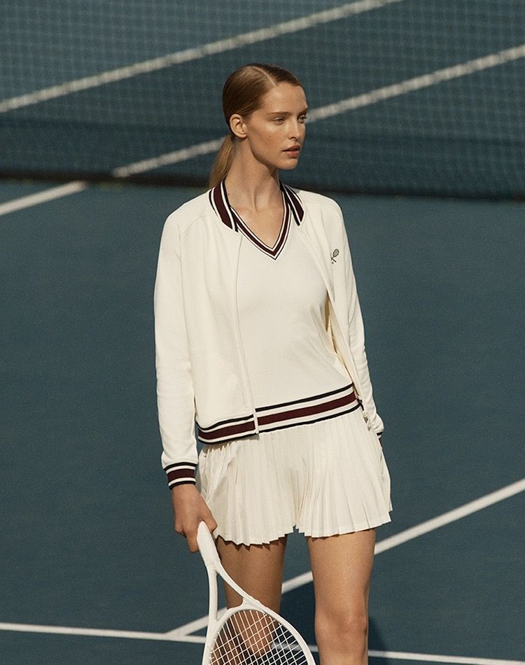 A blonde woman with her hair tied back in a single hair tie, wearing a beige V-neck knit sweater with maroon stripes at the hem, layered with a beige knit cardigan, a beige pleated skirt, and holding a tennis racket.